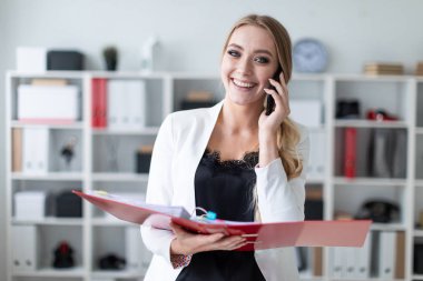 Genç bir kız ofis raf yanında duran, telefonda konuşurken ve belgeler ile bir klasör holding.
