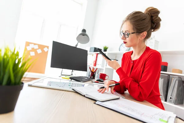 Молодая девушка стоит за столом в офисе с черным маркером и телефоном в руке. Девушка работает с компьютером, блокнотом и магнитной доской . — стоковое фото