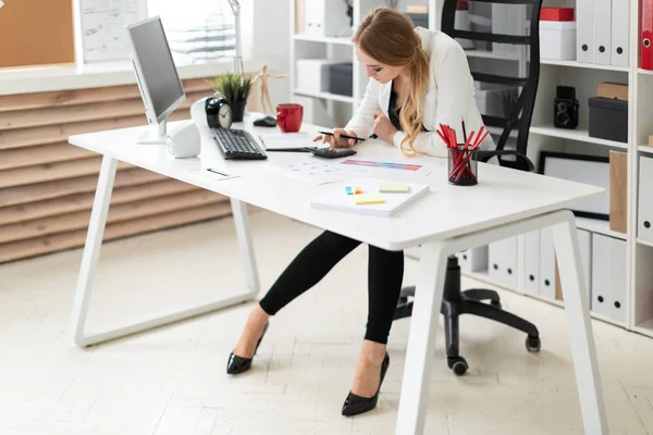 若い女の子は、オフィスのコンピューターのデスクで座っているし、電卓でカウントします。女の子の前にある文書. — ストック写真