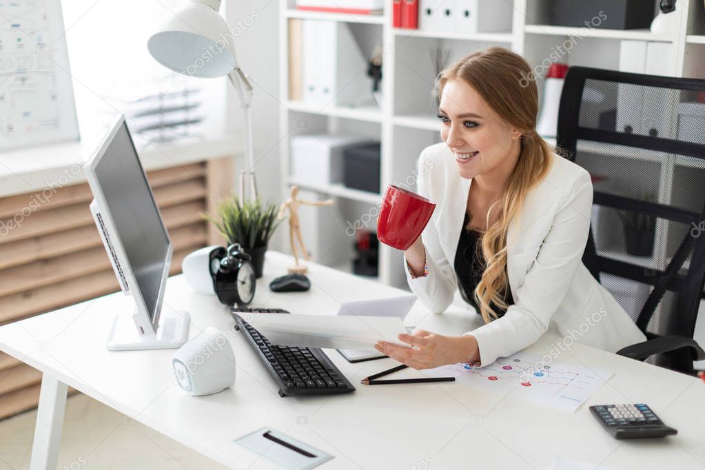 A young girl is sitting at the computer desk in the office and is holding a cup and documents. Before the girl on the table are diagrams.