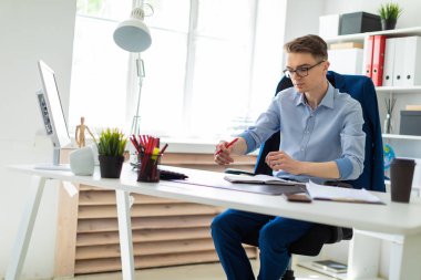 Genç bir adam bir bilgisayar masası ofisinde oturuyor ve bir kalem elinde tutuyor.