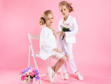 Açık renk elbiseli ikizler çiçek buketleriyle pembe arka planda bir sandalyenin yanında poz veriyorlar..