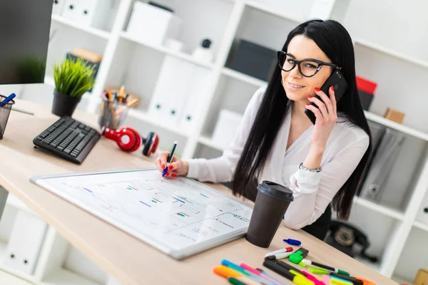 Una joven con gafas se para cerca de una mesa, habla por teléfono y dibuja un marcador en una pizarra magnética . — Foto de Stock