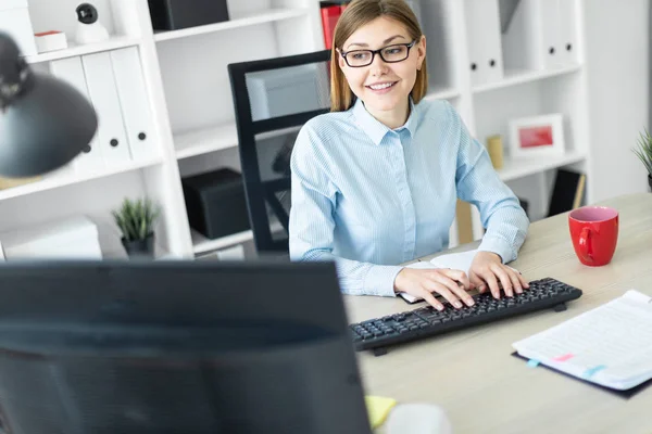 Девушка в очках сидит за столом в офисе, держит карандаш в руке и печатает текст на клавиатуре . — стоковое фото