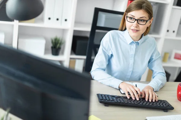 Una joven con gafas se sienta en una mesa en la oficina, sostiene un lápiz en la mano y escribe el texto en el teclado . — Foto de Stock