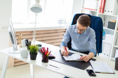 Genç bir adam bir bilgisayar masası ofisinde oturuyor ve bir not defterinde yazıyor.