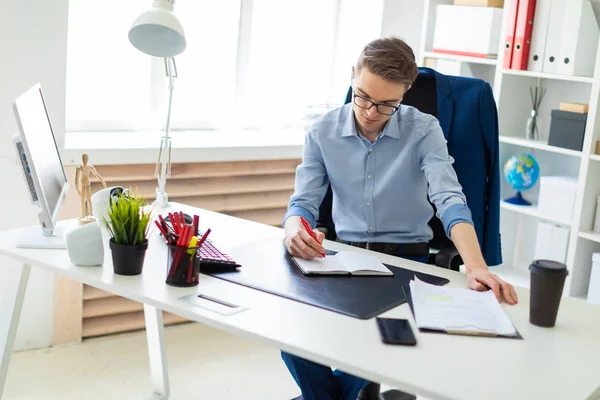 Genç bir adam bir bilgisayar masası ofisinde oturuyor ve bir not defterinde yazıyor.