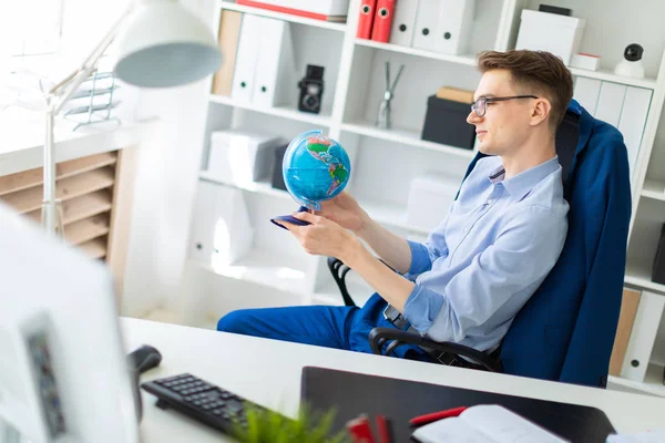 Un jeune homme assis dans le bureau à un bureau d'ordinateur et tient un globe dans ses mains. — Photo