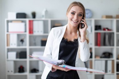 Genç bir kız ofis raf yanında duran, telefonda konuşurken ve belgeler ile bir klasör holding.