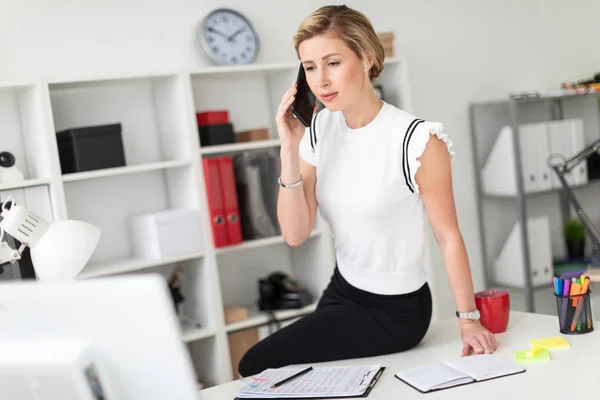 Uma jovem loira está sentada na mesa no escritório e falando ao telefone . — Fotografia de Stock