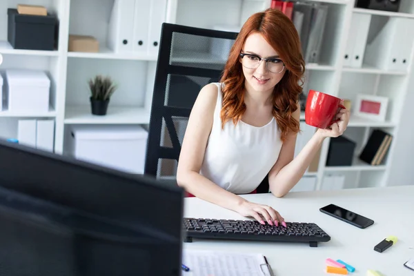 Beautiful young girl sits at the desk in the office, holds a cup in one hand and works at the computer.