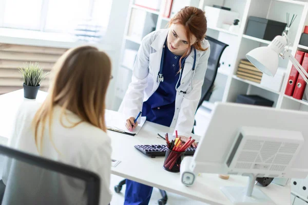 En vacker ung flicka i en vit klädnad är står nära en dator skrivbord på kontoret och kommunicera med samtalspartnern. Flickan gör anteckningar i dokumentet. — Stockfoto