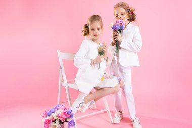 Açık renk elbiseli ikizler çiçek buketleriyle pembe arka planda bir sandalyenin yanında poz veriyorlar..