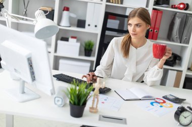Genç bir kız ofiste bir masada oturuyor, kırmızı bir fincan elinde tutan ve klavyede yazarak.