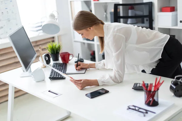 A young girl stands bent near a table in the office. The girl works with a computer, notepad and documents. — Stock Photo, Image