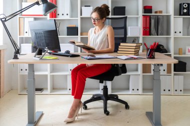 Genç bir kız bilgisayar masasında oturuyor ve elinde açık bir kitap tutuyor..