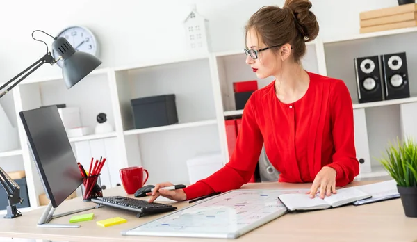 Una joven se sienta en una mesa en la oficina y sostiene un marcador negro en su mano. La chica trabaja con una computadora, bloc de notas y una placa magnética . — Foto de Stock