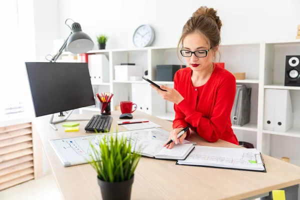 Una joven está de pie en una mesa en la oficina, sosteniendo un marcador negro y un teléfono en la mano. La chica trabaja con una computadora, bloc de notas y una placa magnética . — Foto de Stock