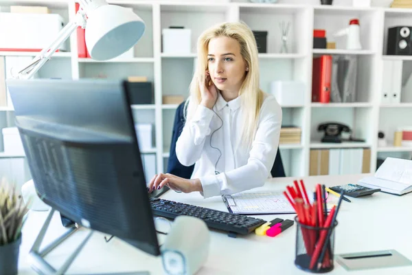 Una joven en la oficina se sienta en una mesa hablando por teléfono a través de un auricular y trabajando en la computadora . — Foto de Stock