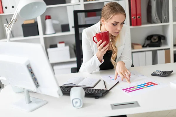 Молодая девушка сидит за компьютерным столом в офисе и держит красную чашку в руке. Перед девушкой на столе диаграммы . — стоковое фото