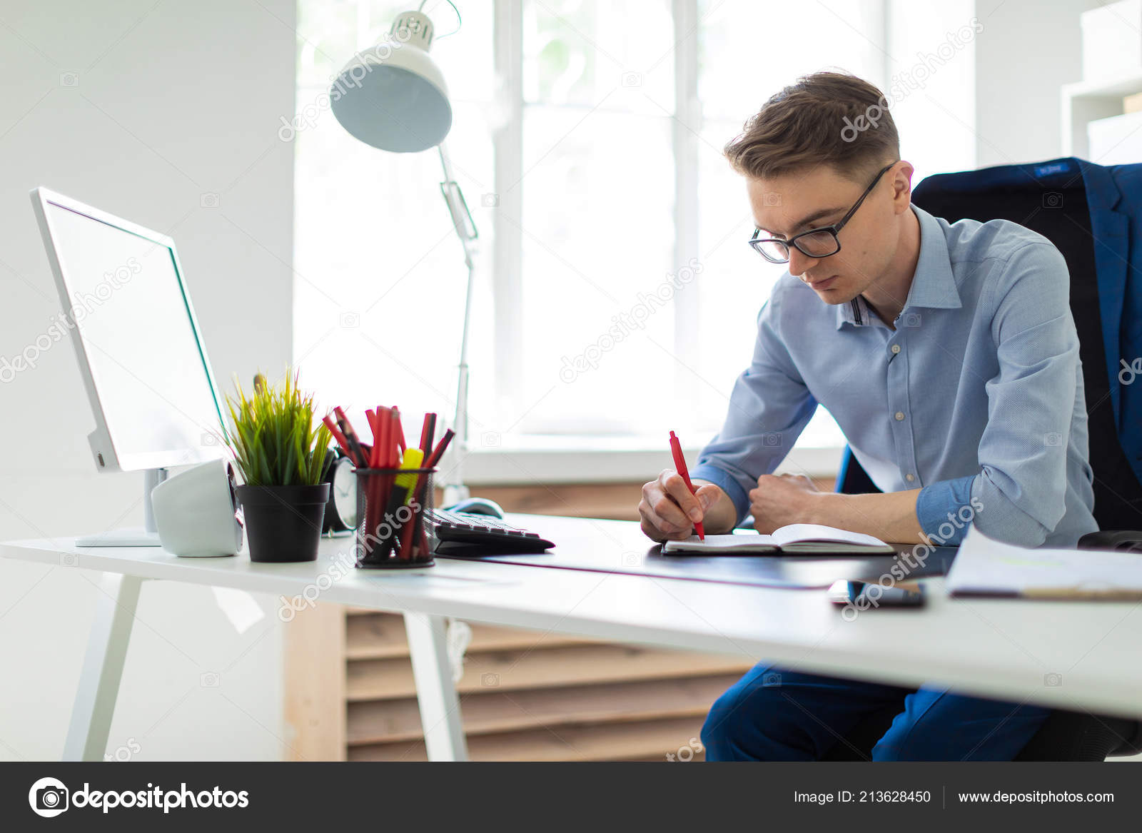 A Young Man Sits In The Office At A Computer Desk And Writes In A