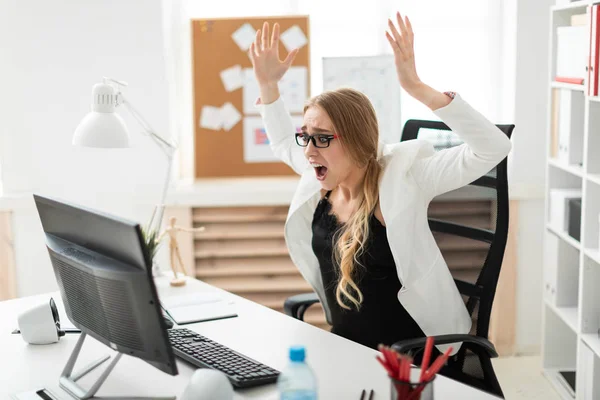 Una joven está sentada en una mesa de la oficina, mirando al monitor, levantando las manos y gritando . — Foto de Stock