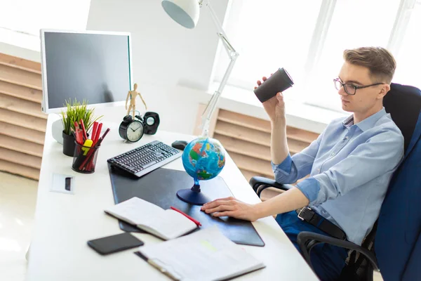 Молодой человек сидит в офисе за компьютерным столом и держит в руке стакан кофе. Молодой человек сталкивается с глобусом. — стоковое фото