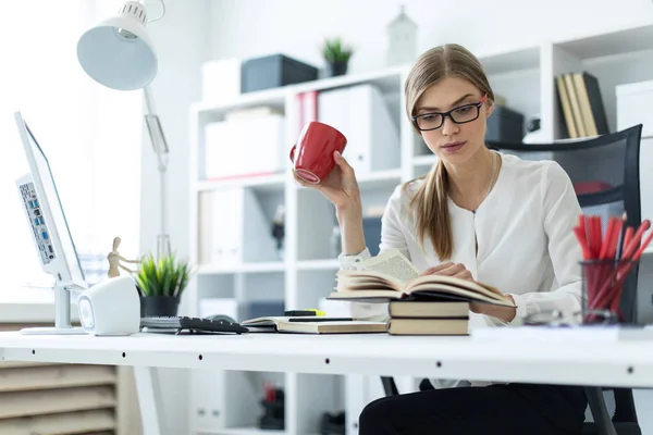 Una joven se sienta en una mesa en la oficina y sostiene una taza roja en su mano y lee un libro . — Foto de Stock