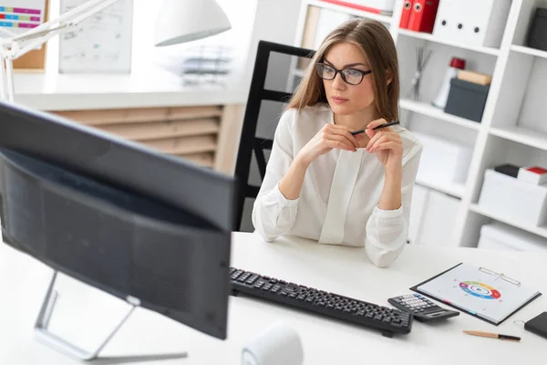 Mladá dívka sedí u stolu v kanceláři, drží tužku v ruce a při pohledu na monitor. — Stock fotografie