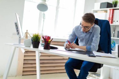 Genç bir adam bir bilgisayar masası ofisinde oturuyor ve bir not defterinde yazıyor.