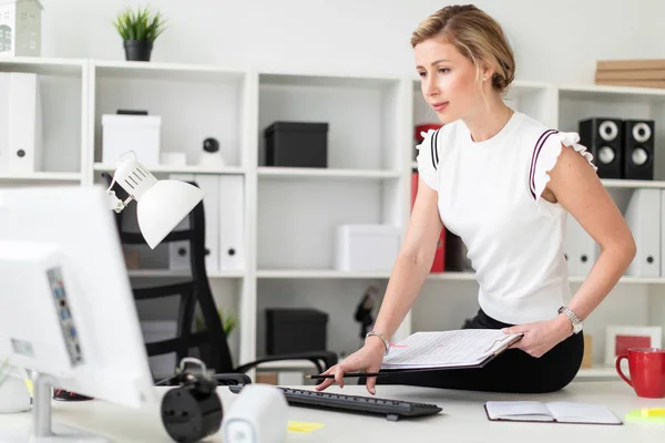 Een jong blond meisje zit op een bureau in het kantoor, houdt van documenten en een potlood in haar hand en drukt op het toetsenbord. — Stockfoto