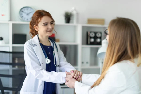 Uma menina bonita com um casaco branco está de pé perto da mesa no escritório e apertando as mãos com o paciente. O paciente agradece ao médico . — Fotografia de Stock