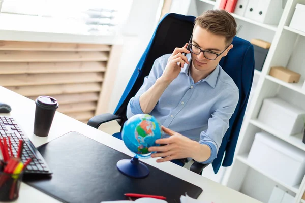 Un giovane uomo si siede in ufficio alla scrivania di un computer, parla al telefono e guarda il globo . — Foto Stock