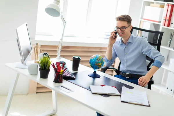 A young man sits in the office at a computer desk, talking on the phone and looking at the globe.