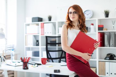 Güzel bir genç kız ofiste masanın üzerine oturdu ve elinde kırmızı bir dosya tutuyordu..