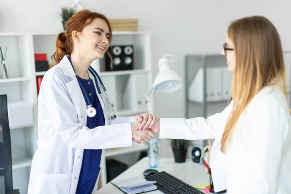 Uma menina bonita com um casaco branco está de pé perto da mesa no escritório e apertando as mãos com o paciente. O paciente agradece ao médico . — Fotografia de Stock