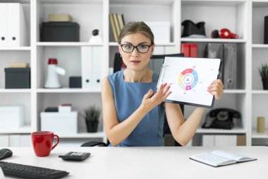 Genç bir kız ofiste bir masada oturur ve bir kalem ve bir diyagram bir belgeyle tutar.