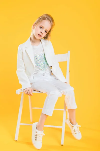 Ein junges Mädchen in weißen Kleidern sitzt auf einem Stuhl vor gelbem Hintergrund. — Stockfoto