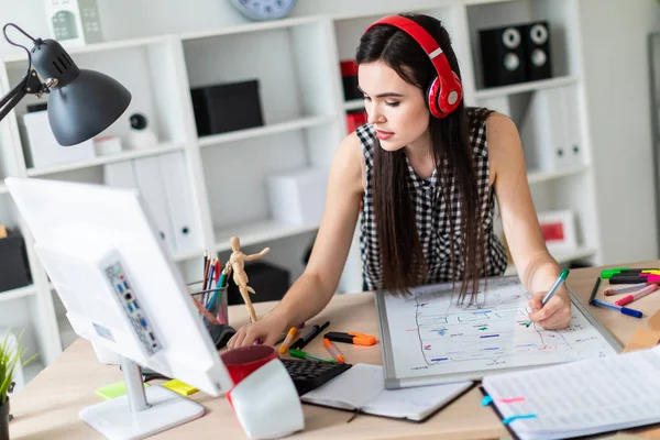 Mladá dívka stojí u stolu, zelená značka drží v ruce a pracuje s počítačem a Magnetická tabule. Na hlavě dívka, která nosí sluchátka. — Stock fotografie