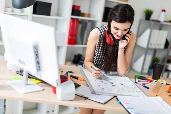 Uma jovem está parada perto de uma mesa, segurando um marcador verde na mão e falando ao telefone. No pescoço, os fones de ouvido das meninas pendurados . — Fotografia de Stock