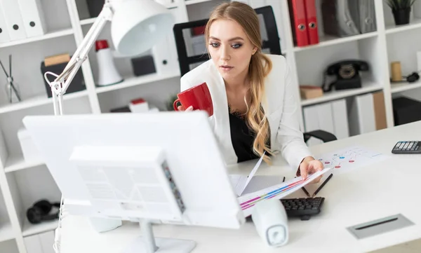 Молодая девушка сидит за компьютерным столом в офисе и держит в руках чашку и документы. Перед девушкой на столе диаграммы . — стоковое фото