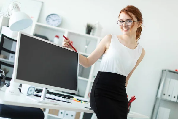 A beautiful young girl in the office sat down on the table and was holding a red pencil in her hand. Next to the girl is a monitor.