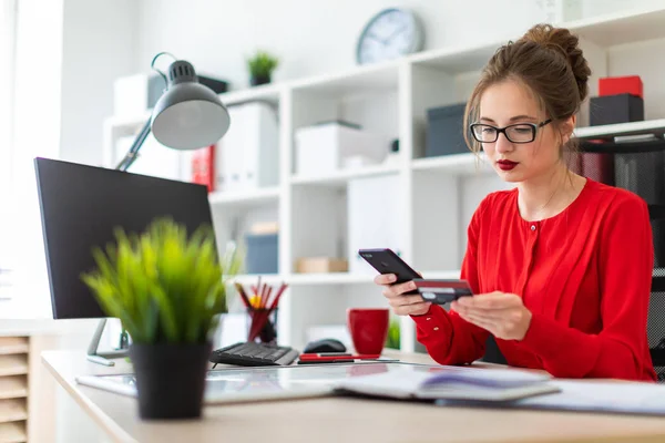 Una joven está sentada en el escritorio de la oficina, con una tarjeta bancaria y un teléfono en la mano. . — Foto de Stock