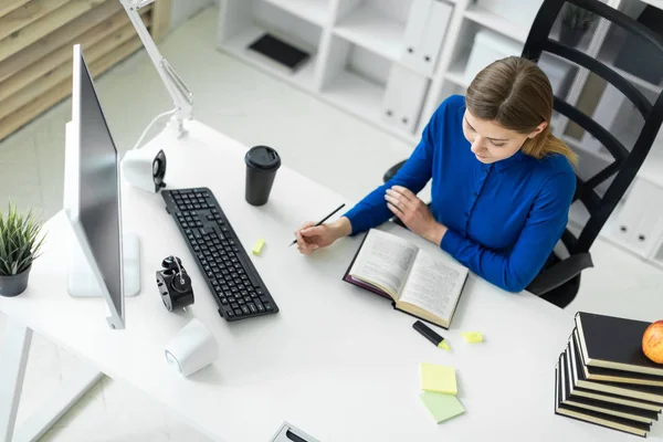 Una joven se sienta en un escritorio de computadora y sostiene un lápiz en su mano. Antes de que la chica mienta un libro abierto . — Foto de Stock