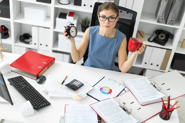 Молодая девушка сидит за столом в своем офисе, держа будильник и красную чашку . — стоковое фото