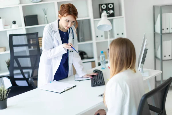 En vacker ung flicka i en vit klädnad är står nära en dator skrivbord på kontoret och kommunicera med samtalspartnern. Flickan gör anteckningar i dokumentet. — Stockfoto