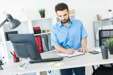 Genç adam bir bilgisayar danışma belgeleri, diyagramları ve telefon ile ofiste çalışır.