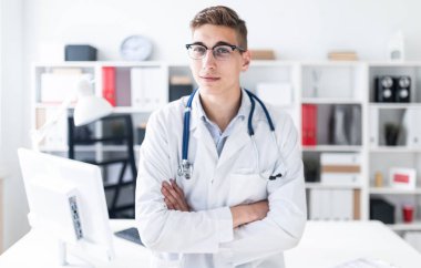 Önlük üzerinde boyun ayakta stetoskop ile genç doktor ofisinde çapraz kolları olan