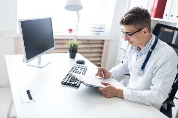 年轻的医生拿着剪贴板 一边坐在办公室里拿着电脑的办公桌前检查病史 — 图库照片