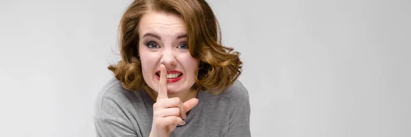 Encantadora joven con una camiseta gris sobre un fondo gris. La chica inclinó su dedo hacia sus labios — Foto de Stock